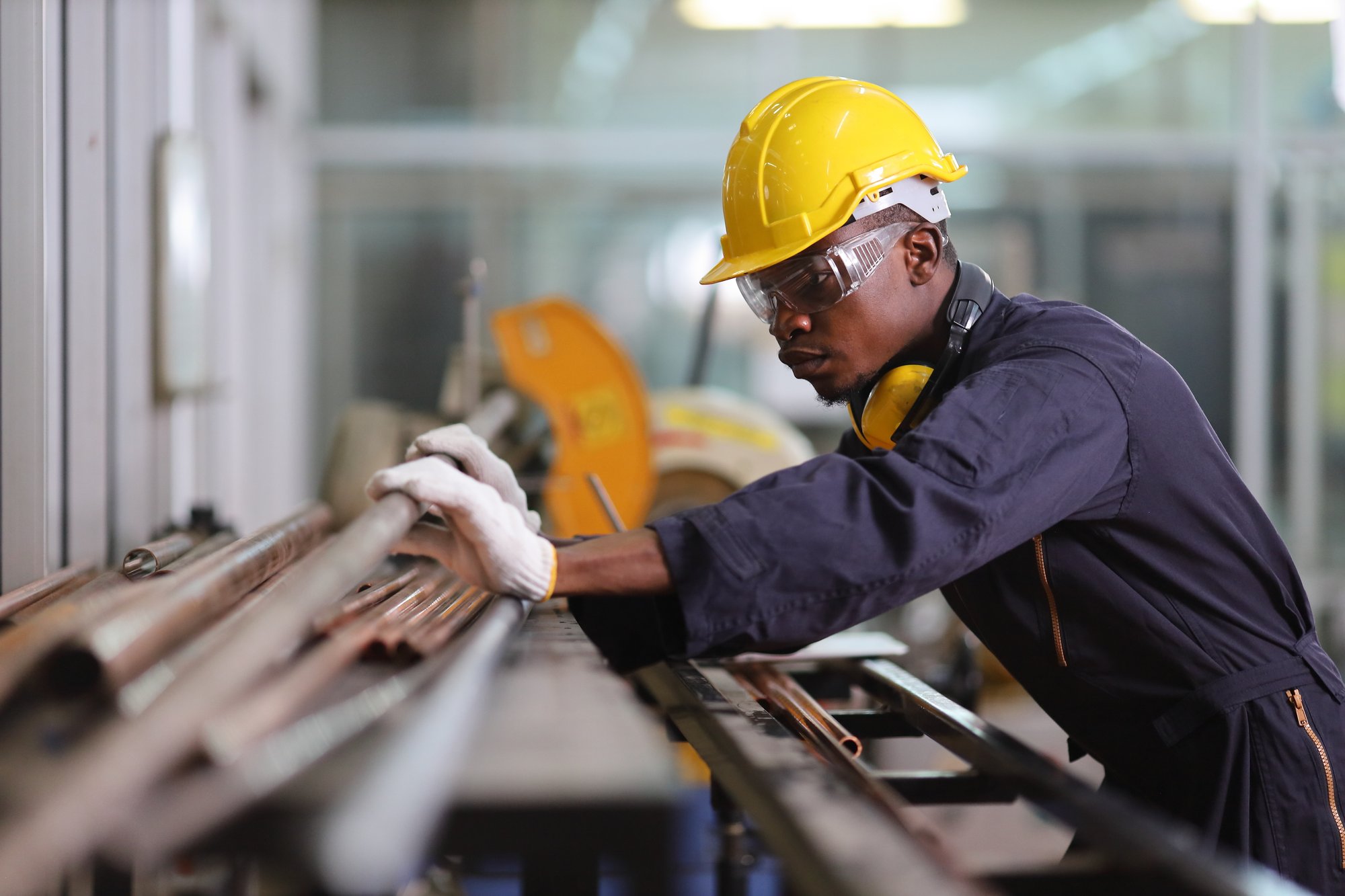 A worker in a yellow hard hat and protective goggles is inspecting metal pipes on an assembly line, seamlessly integrating AI in manufacturing. He dons gloves and a dark work uniform, with industrial machinery humming in the background.
