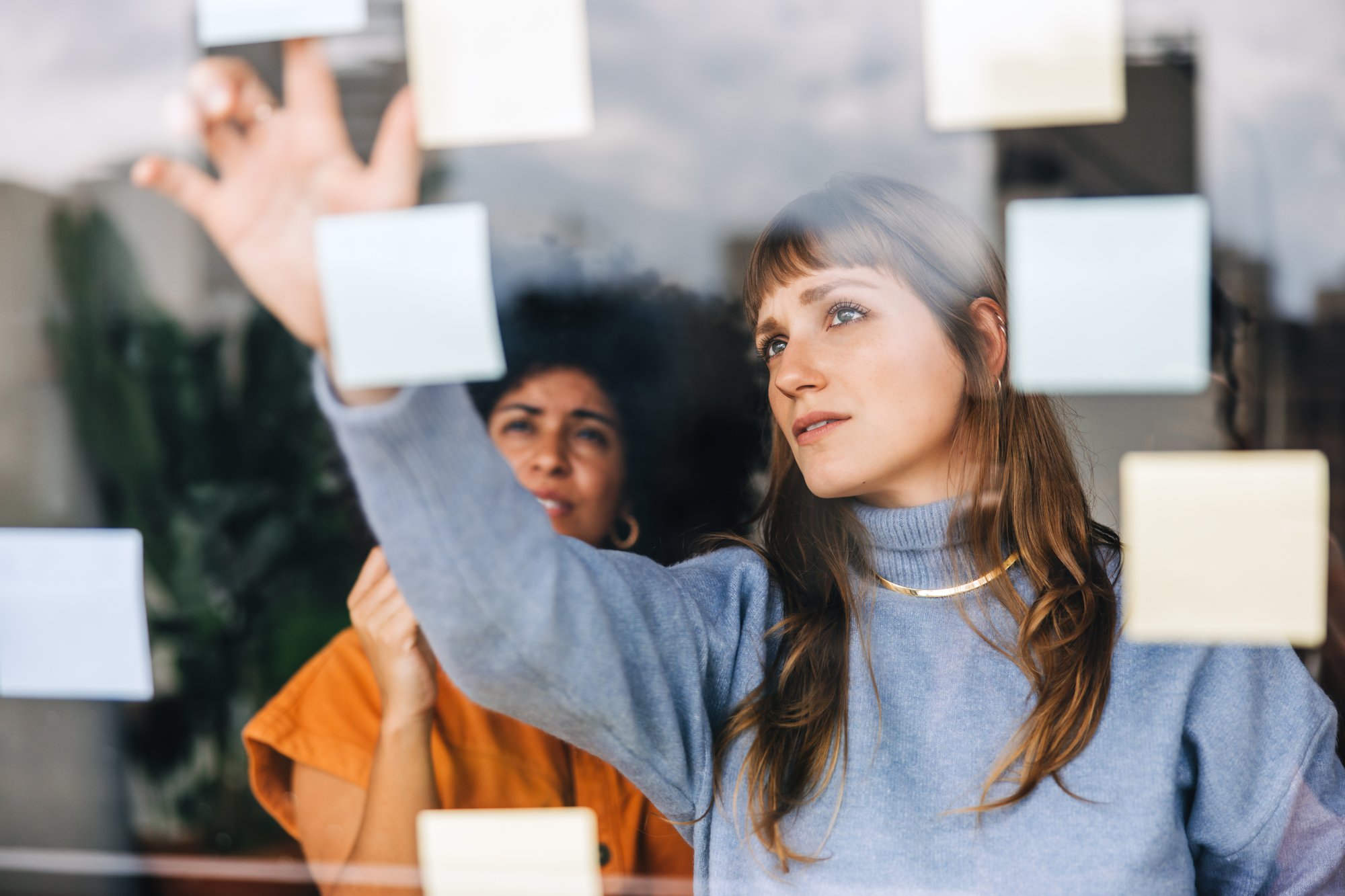 Two women are seen through a glass wall covered with sticky notes, brainstorming ideas. One in a blue sweater reaches out to place or adjust a note, while the other, in orange, looks on—perhaps discussing AI in private equity.